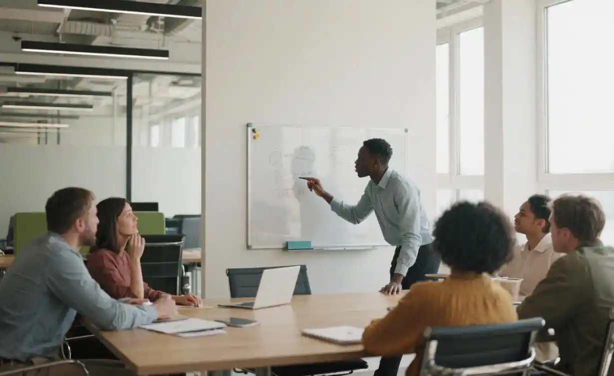 a young Black man leaning forward to share an idea on a whiteboard, colleagues listening with interest